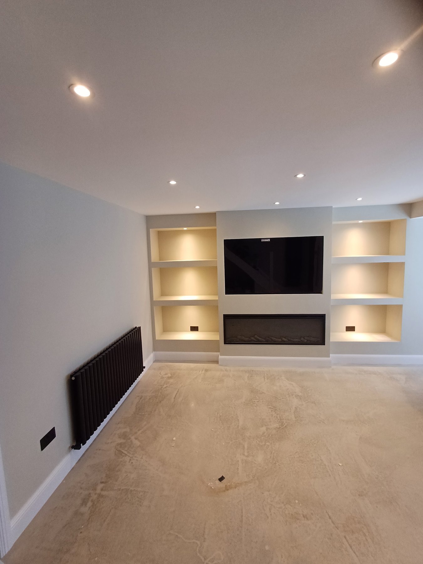 A long room freshly painted, with a media wall, lit alcove shelves and a pale carpet